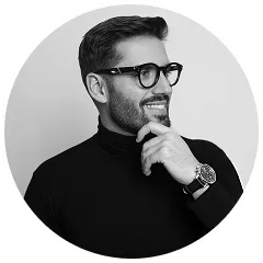 Black and white portrait of a smiling man with glasses and a watch, wearing a black turtleneck, looking to the side with his hand on his chin.