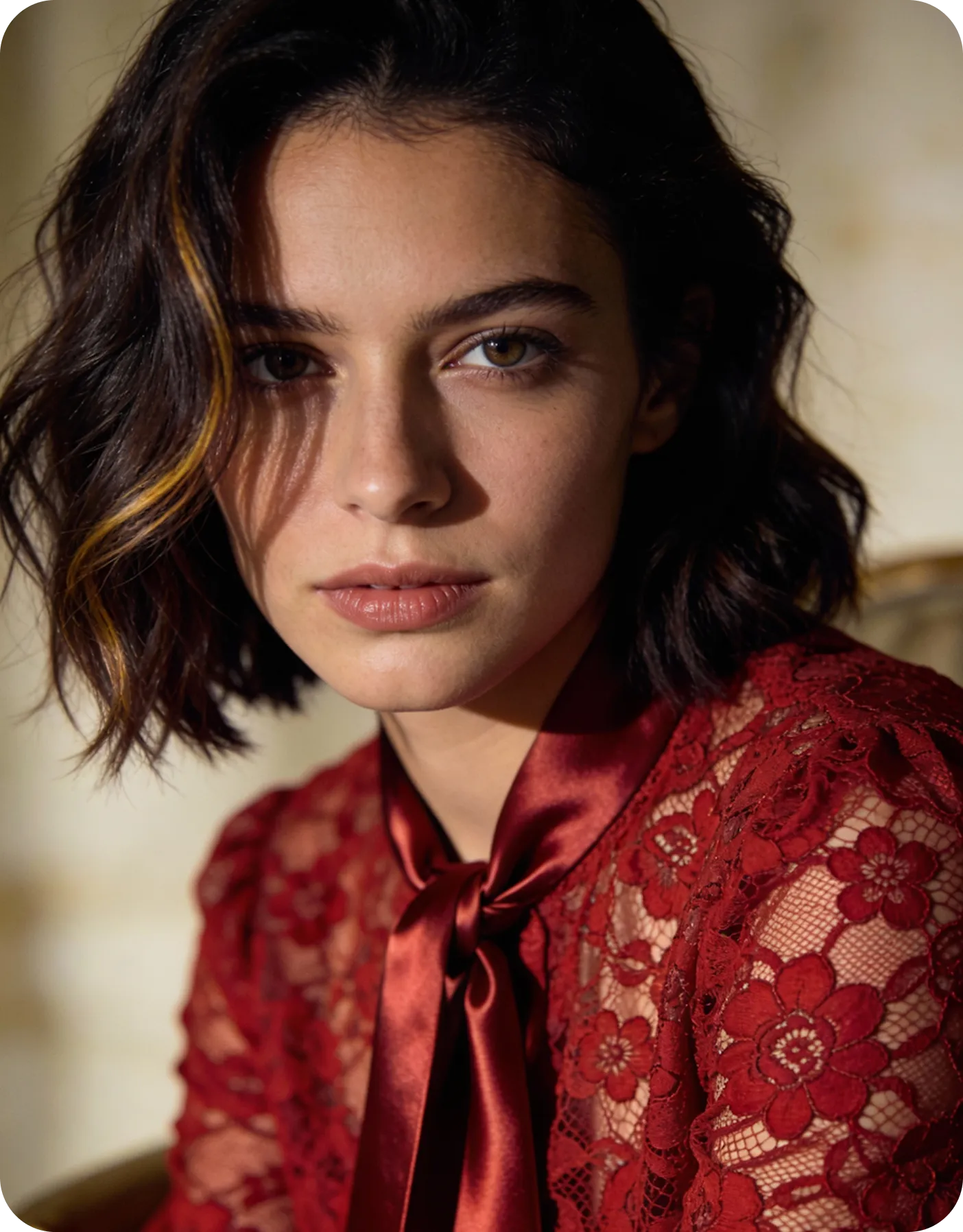Young woman with shoulder-length dark hair and hazel eyes wearing a red lace top with a satin bow tie collar.