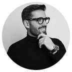 Smiling man with glasses and a beard wearing a black turtleneck, looking to the side with hand near chin.
